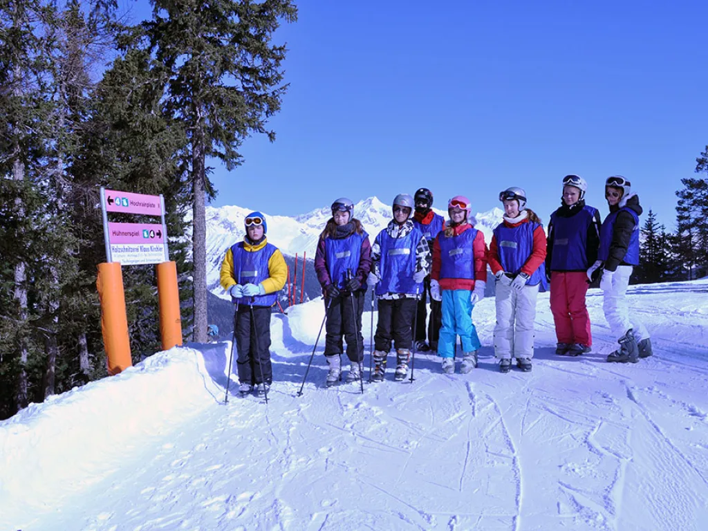 Skigruppe steht neben einer Pistentafel vor der Bergkulisse.